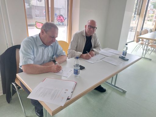 Zwei Männer sitzen an einem Tisch, schreiben Notizen auf Papier und haben Wasserflaschen dabei.